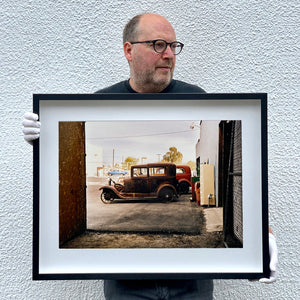 Black framed photograph held by Richard Heeps. The side view of two vintage early Ford Motor vehicles parked in a yard. 