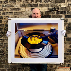 Photograph held by photographer Richard Heeps. A black vinyl with a yellow label sits high on the spindle of a deck under the overarm.