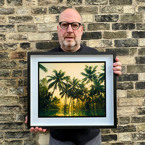 Black framed photograph held by photographer Richard Heeps. Green palms trees against a warm golden sky, and reflected in a pool below.