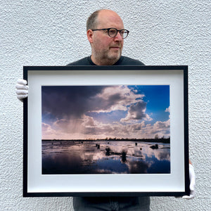 Black framed photograph held by photographer Richard Heeps. Fenland expanse with water and tufts of grass and an expansive fenland sky, blue with white and grey clouds reflected in the water below.