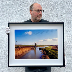 Black framed photograph held by photographer Richard Heeps. Fenland waterway with a weight drain up in the middle. Fenland sits either side and there is a blue sky.