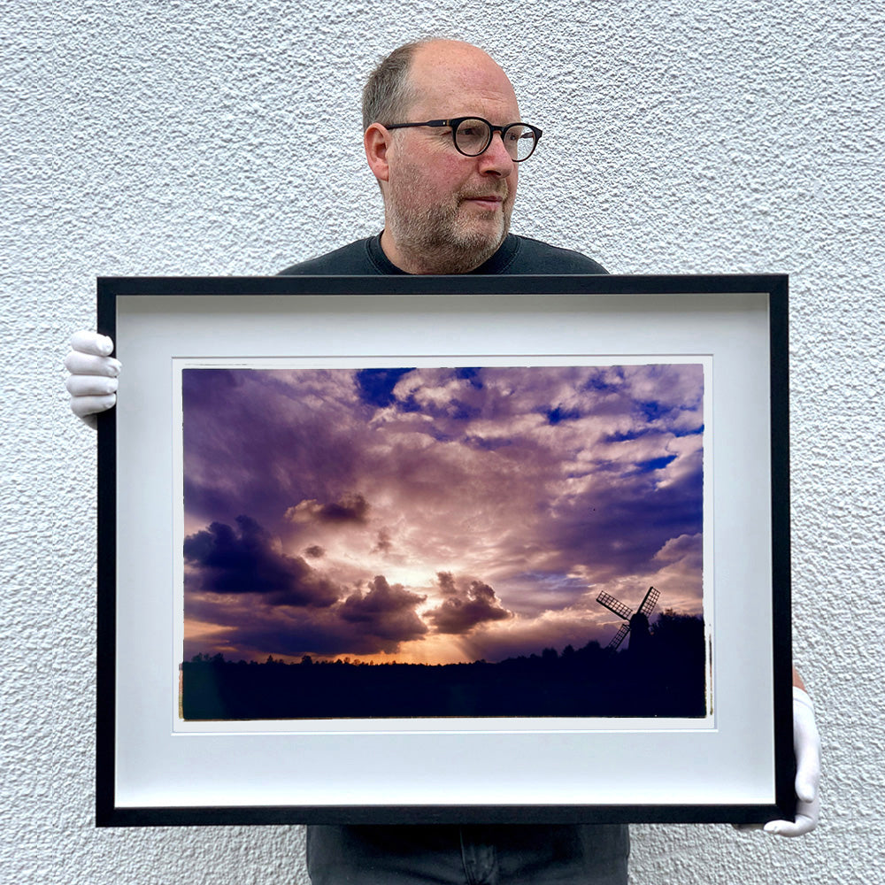 Black framed photograph held by photographer Richard Heeps. A silhouette of a tree lined fen flat land with a windpump sitting on the right hand side. The sky is cloudy and bathed in golden dusk light.