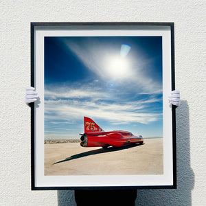 Photograph held by photographer Richard Heeps. A red drag car with a 75 written on its fin sits on a salt plain the front facing away towards the right. A blue cloudy sky is overhead.