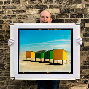 Photograph held by photographer Richard Heeps. Yellow and green beach lockers stand on their wooden legs on the clear sandy beach.