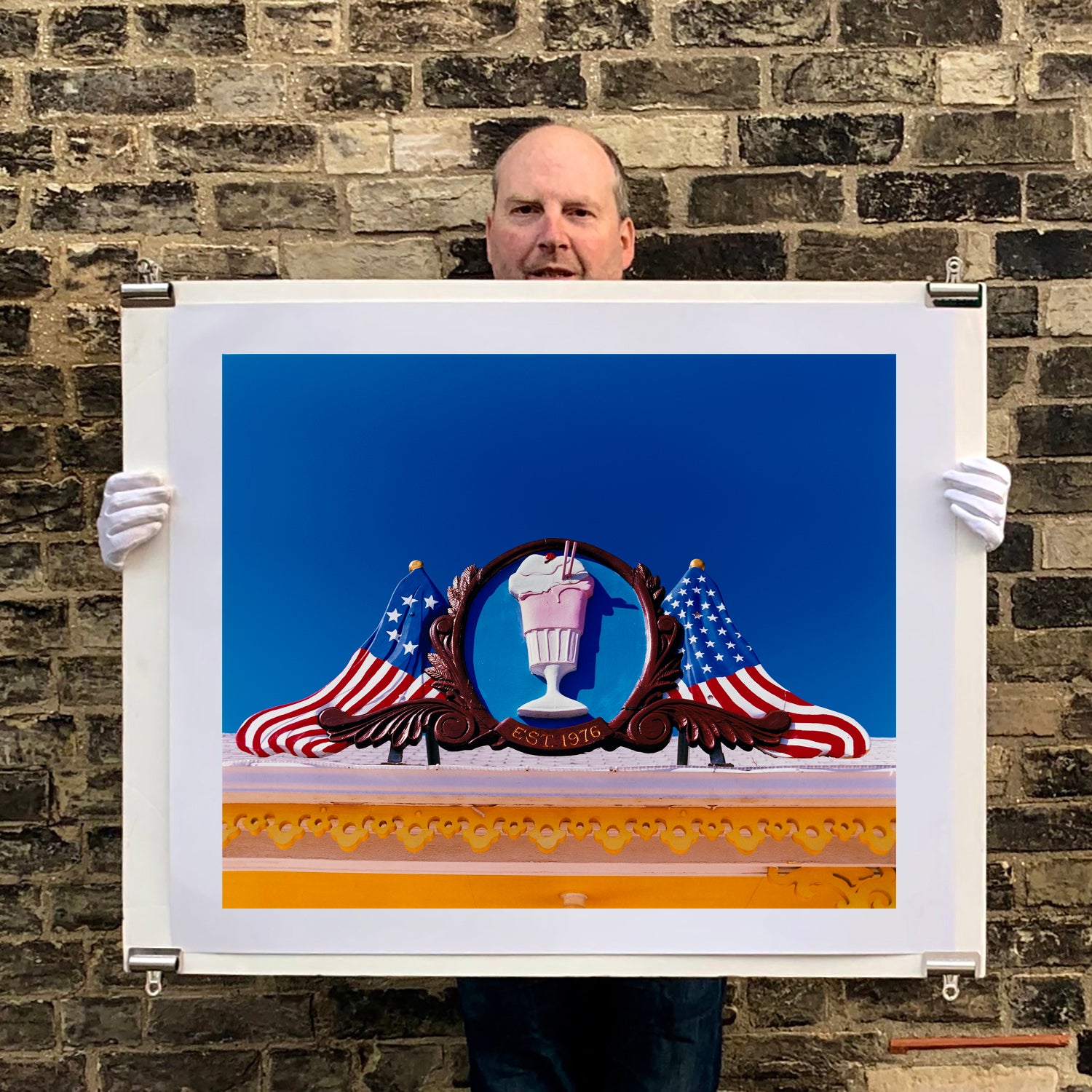 Photograph held by photographer Richard Heeps. A 3D shape milkshake parlour sign which has a pink milkshake with a white top, cherry and straws, surrounded by a wooden type shield, and on either side the look of draped American flags. This is set against a blue sky.