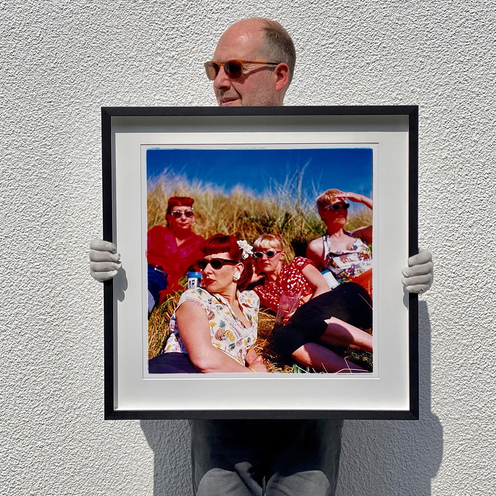 Black framed photograph held by photographer Richard Heeps. The photo is of four women dressed in a retro style all wearing sunglasses on a sunny day lounging on a hill with high grass.