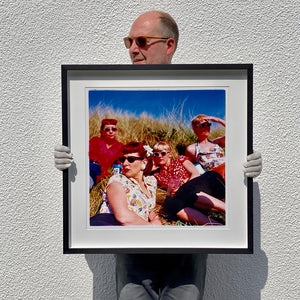Black framed photograph held by photographer Richard Heeps. The photo is of four women dressed in a retro style all wearing sunglasses on a sunny day lounging on a hill with high grass.