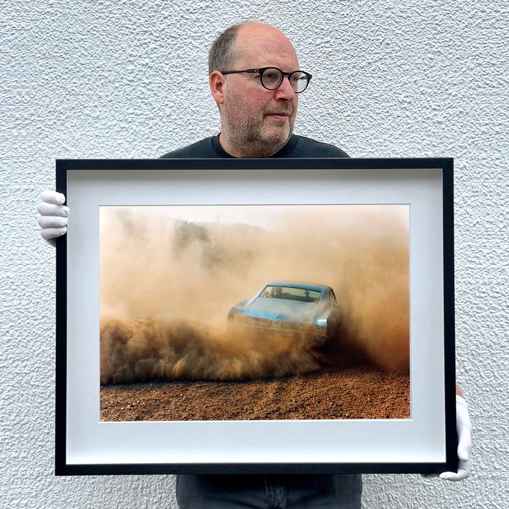 Black framed photograph held by photographer Richard Heeps. A back view of a light blue Buick car moving and slightly obscured by the dust clouds which it has created.