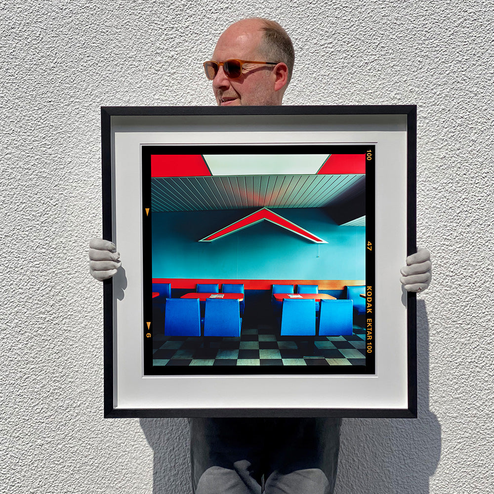 Black framed photograph held by Richard Heeps. This photograph captures the inside of a Wimpy Restaurant in Norfolk. There is bright blue seats and red tables. The walls are blue and there is a big red chevron light attached to the wall. 