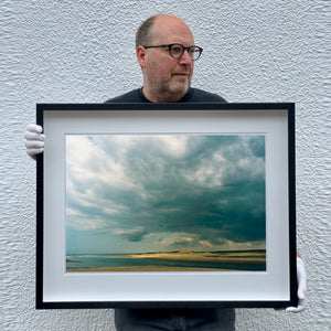Photograph held by photographer Richard Heeps. The photo depicts a dark rolling sky sitting over a Norfolk headland, pouring different lights over the sea, sand and dunes below.