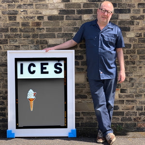 Photograph by Richard Heeps.  Richard Heeps holds a white framed print. At the top of the print, black letters spell out ICES and below is depicted a 99 icecream cone sitting left of centre against a grey coloured background.  
