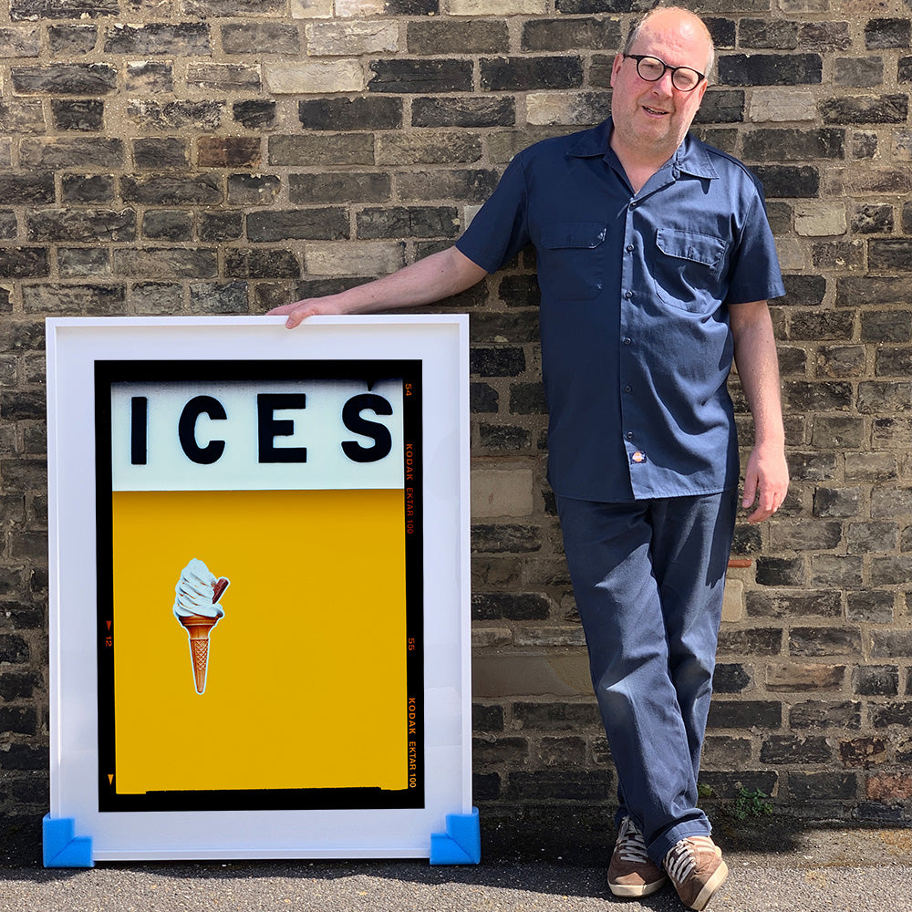 Photograph by Richard Heeps.  Richard Heeps holds a white framed print. At the top of the print, black letters spell out ICES and below is depicted a 99 icecream cone sitting left of centre against a mustard yellow coloured background.  
