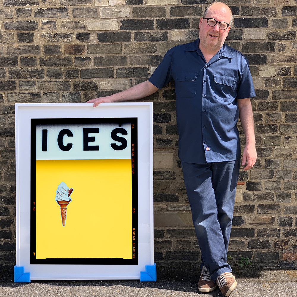 Photograph by Richard Heeps.  Richard Heeps holds a white framed print. At the top of the print, black letters spell out ICES and below is depicted a 99 icecream cone sitting left of centre against a sherbert yellow coloured background.  