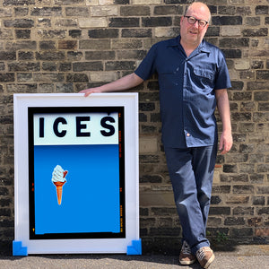 Photograph by Richard Heeps.  Richard Heeps holds a white framed print. At the top of the print, black letters spell out ICES and below is depicted a 99 icecream cone sitting left of centre against a sky blue coloured background.  