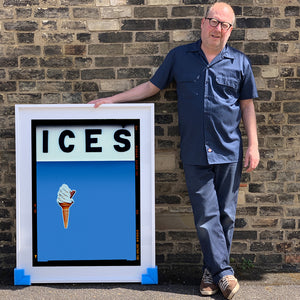 Photograph by Richard Heeps.  Richard Heeps holds a white framed print. At the top of the print, black letters spell out ICES and below is depicted a 99 icecream cone sitting left of centre against a baby blue coloured background.  