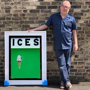 Photograph by Richard Heeps.  Richard Heeps holds a white framed print. At the top of the print, black letters spell out ICES and below is depicted a 99 icecream cone sitting left of centre against a green coloured background.  