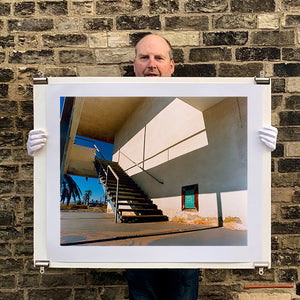 Photograph held by photographer Richard Heeps. The photograph has a metal staircase on the outside of a cream colour motel. The staircase has a ceiling but no sides so leads to blue sky. Behind the building is a blue sky and palm trees of the Californian Desert.