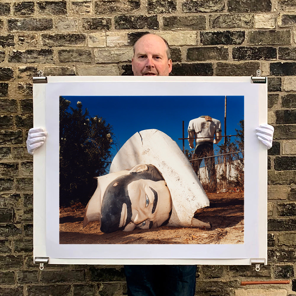 Photograph held by photographer Richard Heeps. In the foreground of the photograph is a broken off head of a giant fibreglass sculpture of a cowboy (a Muffler Man). In the background set in a deep blue sky is the muffler man's headless torso.