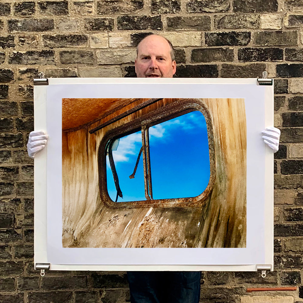 Photograph held by photographer Richard Heeps. The interior of an eroded trailer, looking through the broken window to a deep blue sky.