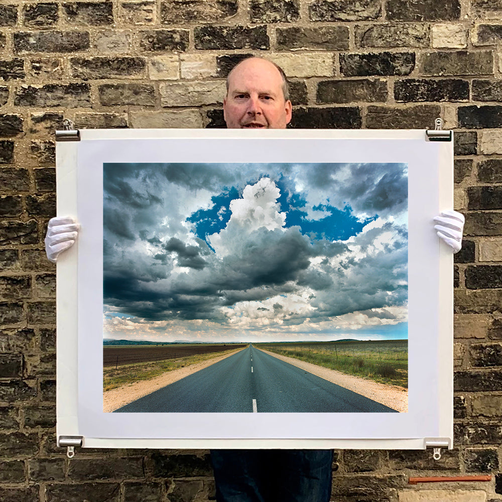 Photograph held by photographer Richard Heeps. A photograph looking along a long straight road into the distance, it is edged either side with sand and then prairie land. The vast, cloudy sky sits above.