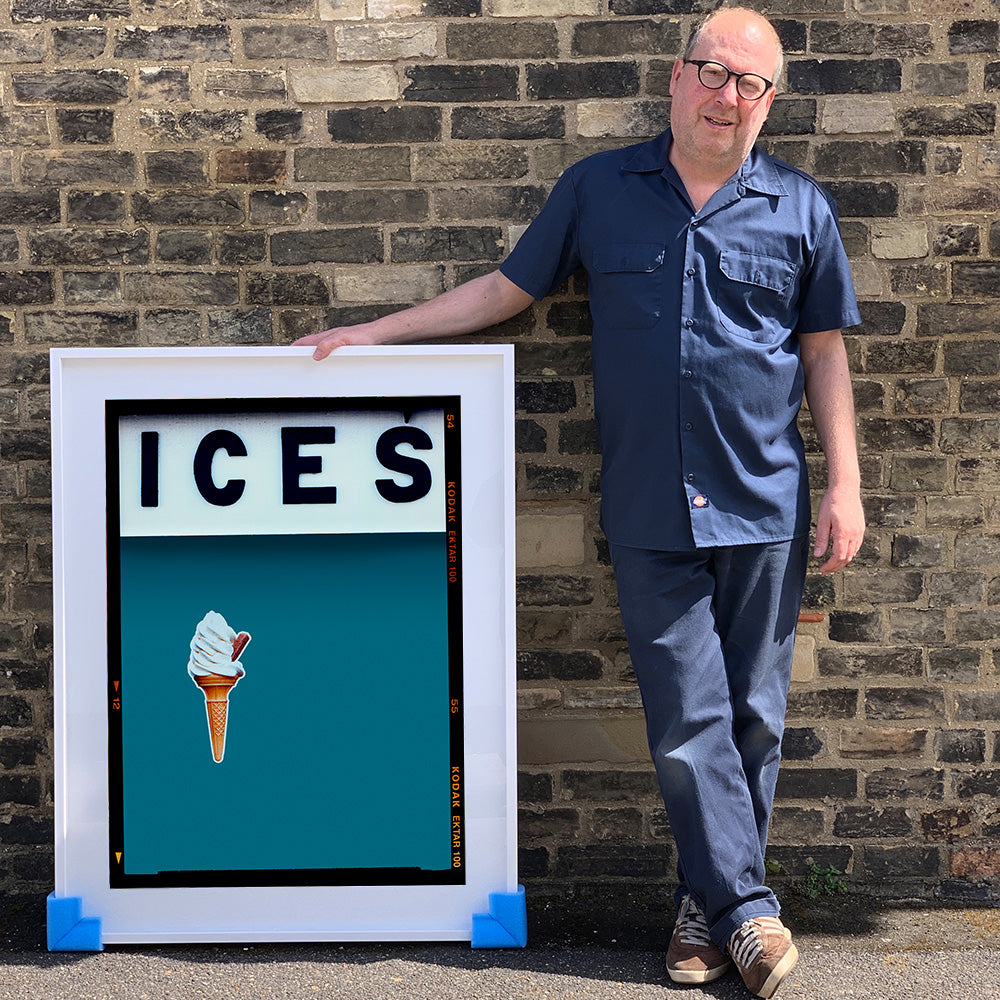 White framed photograph held by photographer Richard Heeps.  At the top black letters spell out ICES and below is depicted a 99 icecream cone sitting left of centre against a blue teal coloured background.  