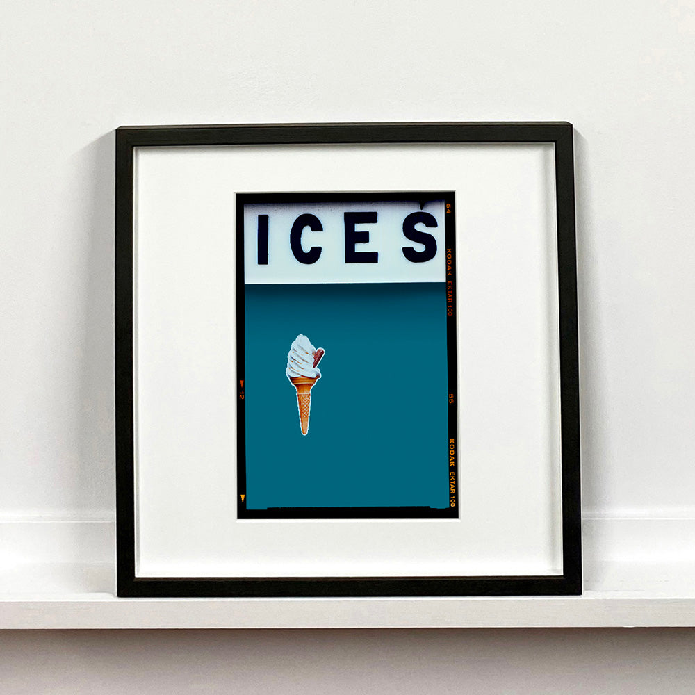 Black framed photograph by Richard Heeps.  At the top black letters spell out ICES and below is depicted a 99 icecream cone sitting left of centre against a blue teal coloured background.  