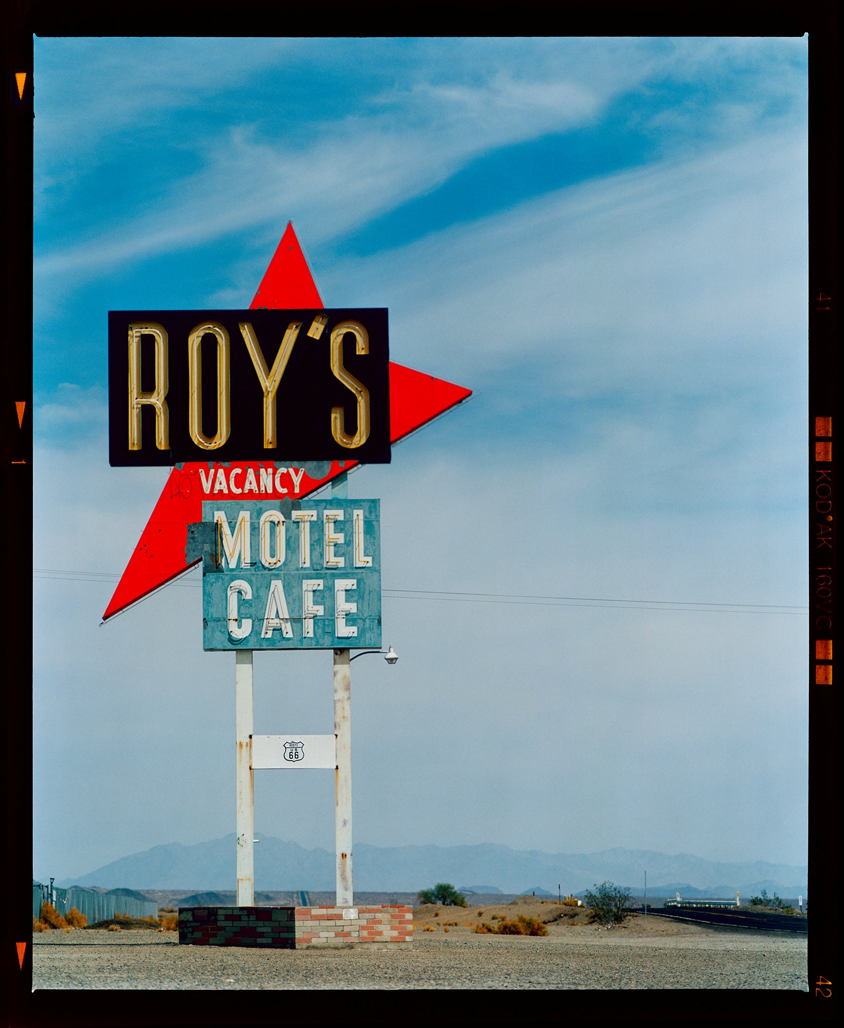 Photograph by Richard Heeps edged by film rebate. A roadside sign on Route 66 in America. The word ROY'S appears in a black sign with a bit red arrow pointing to the left ground, below this VACANCY and on a green square the words MOTEL and CAFE.