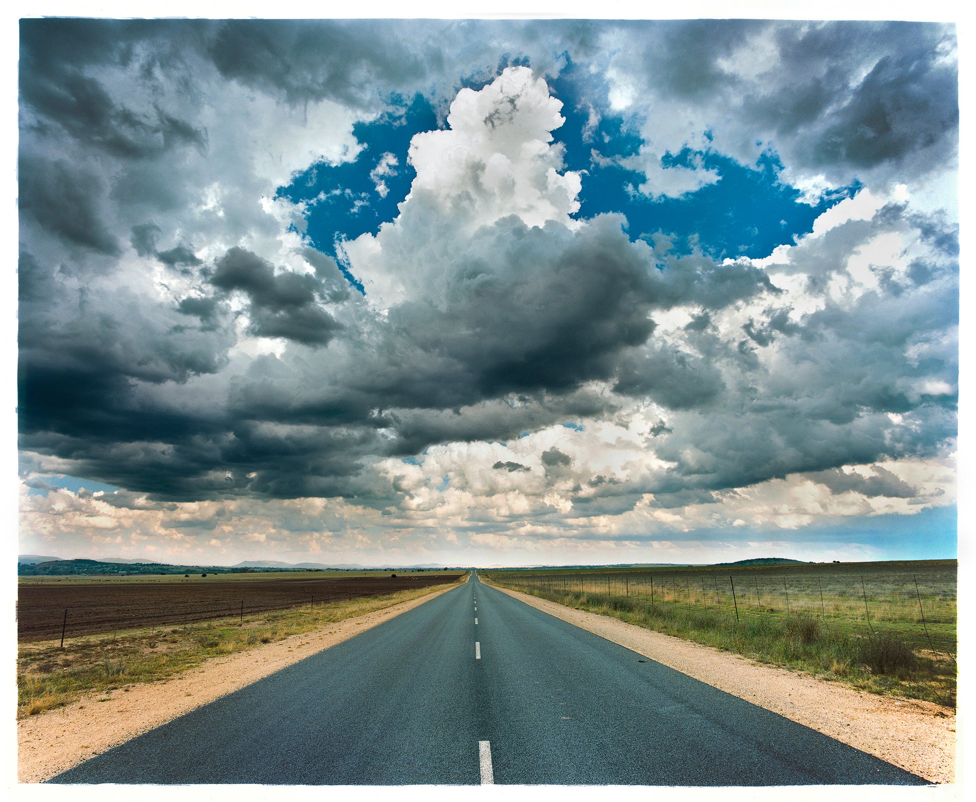 Photography by Richard Heeps. A photograph looking along a long straight road into the distance, it is edged either side with sand and then prairie land. The vast, cloudy sky sits above.