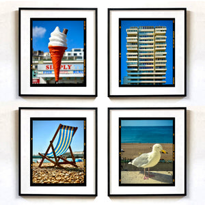 Four black framed wall art prints arranged in a 2x2 grid on a white wall. The prints show a soft-serve ice cream cone, a tall apartment building, a striped beach chair on pebbles, and a seagull by the beach photographs by Richard Heeps