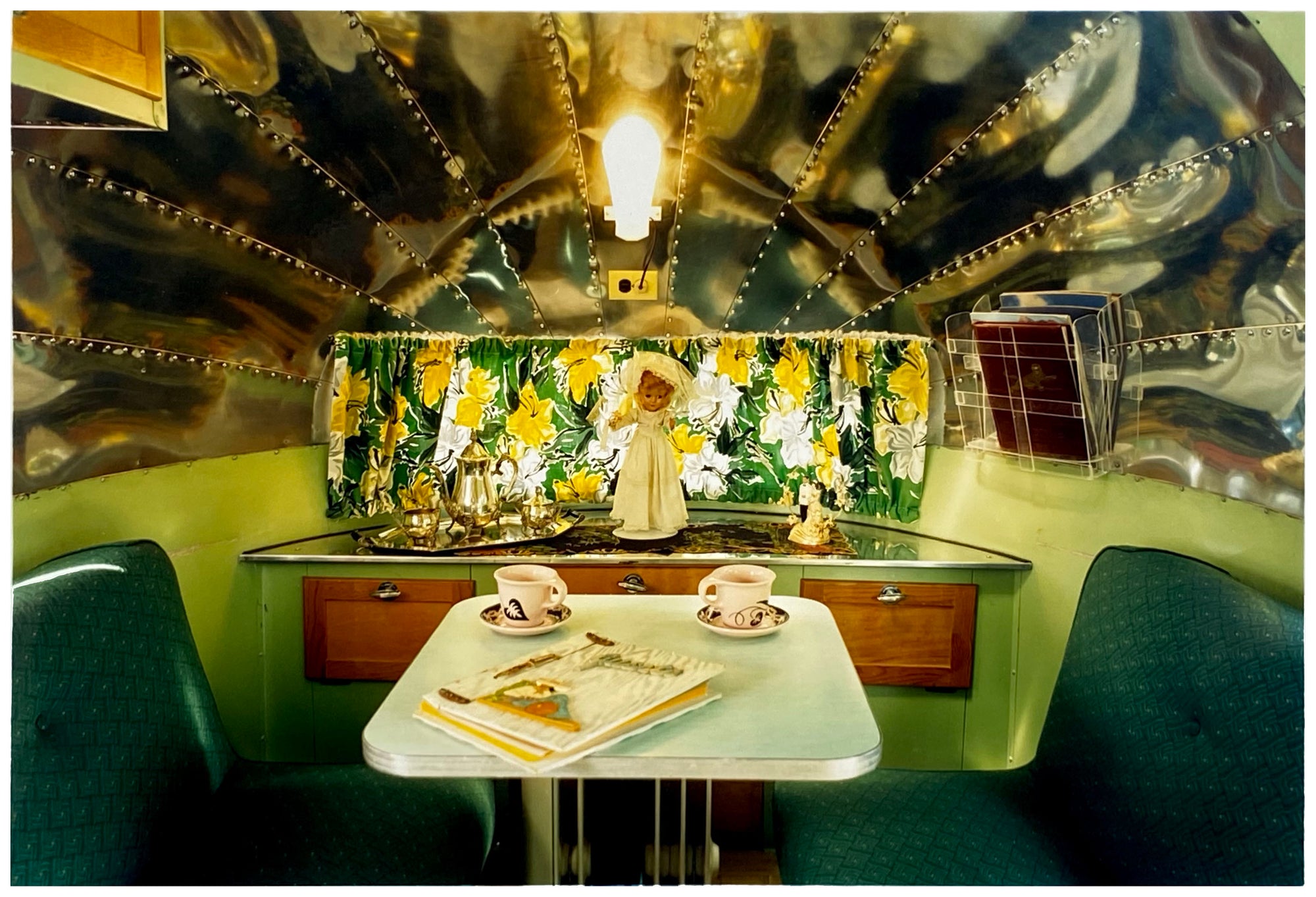 Photograph by Richard Heeps. Inside a trailer, there is a fixed table and chairs with two tea cups and saucers on the table. Behind on the shelf is a tea set and a doll dressed as a bride.