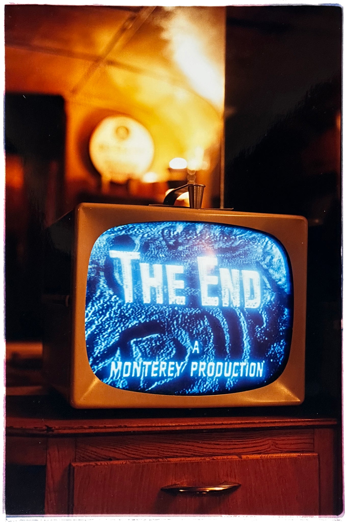 Photograph by Richard Heeps. A retro small brown television sits on a wooden table displaying The End on a black and white film which shows as blue and white. In the background of the television is a warm light.