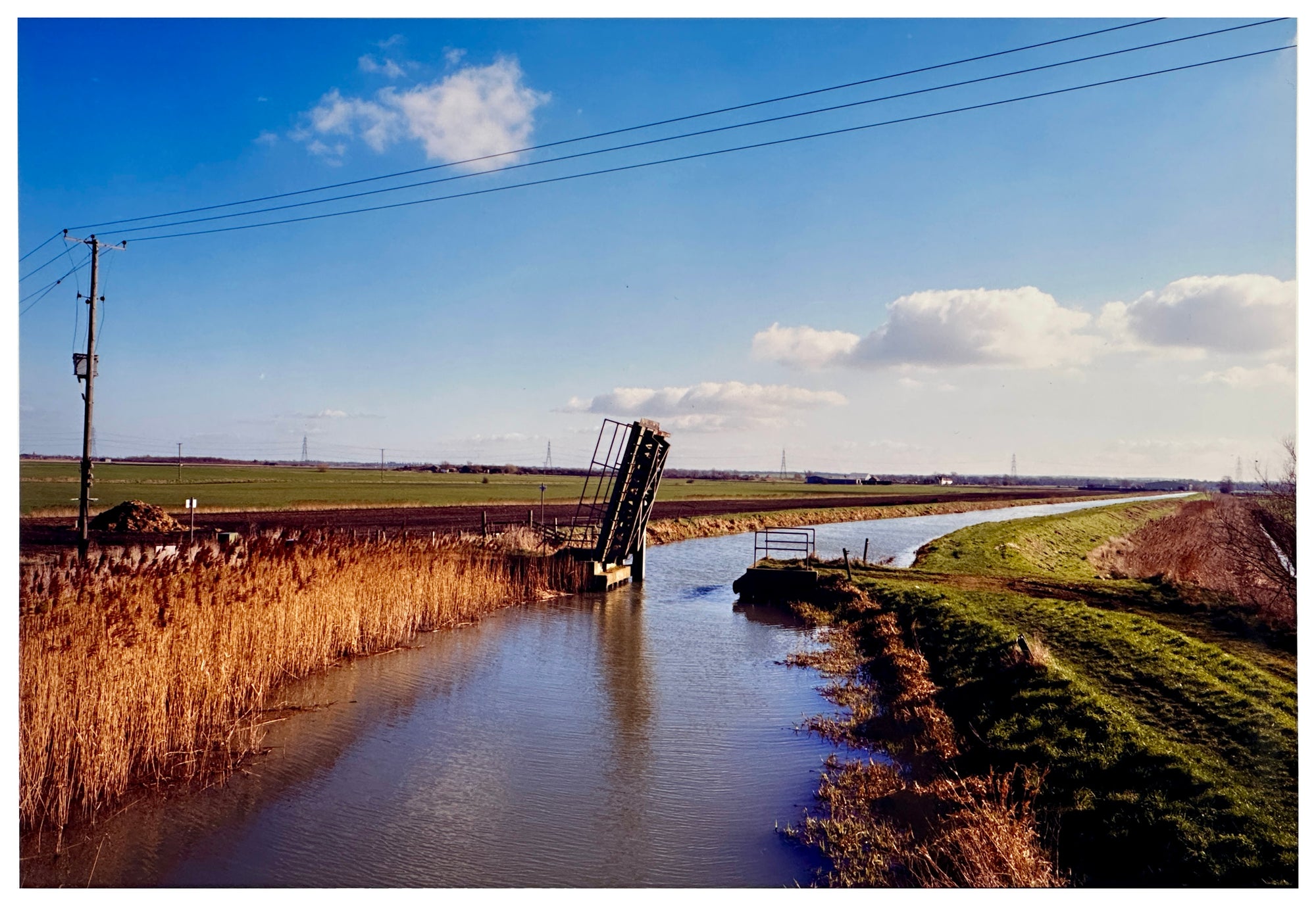 Photograph by Richard Heeps. Fenland waterway with a weight drain up in the middle. Fenland sits either side and there is a blue sky.