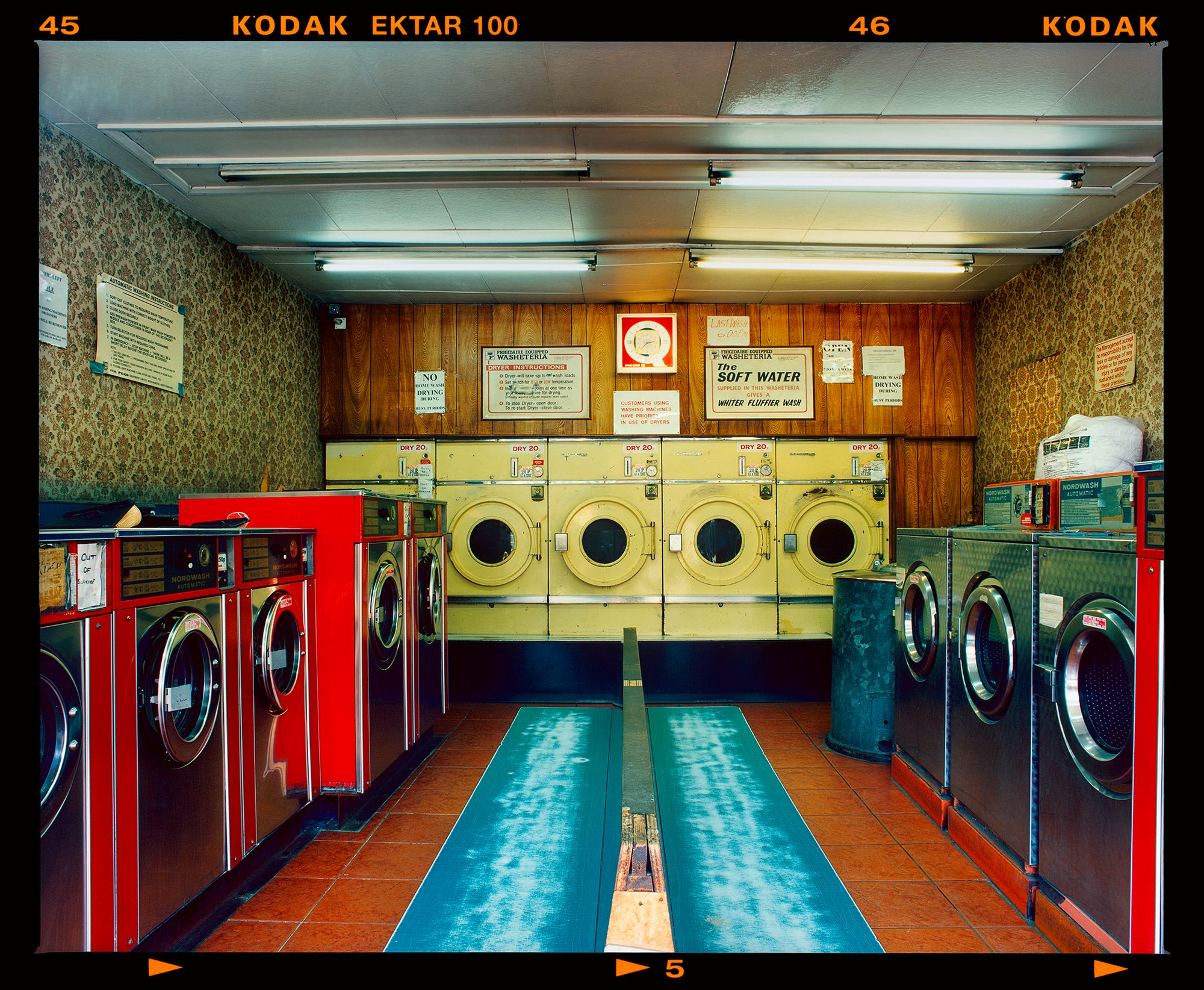 Photograph by Richard Heeps. A laundrette with washing machines on each wall and a double sided seat in the middle.