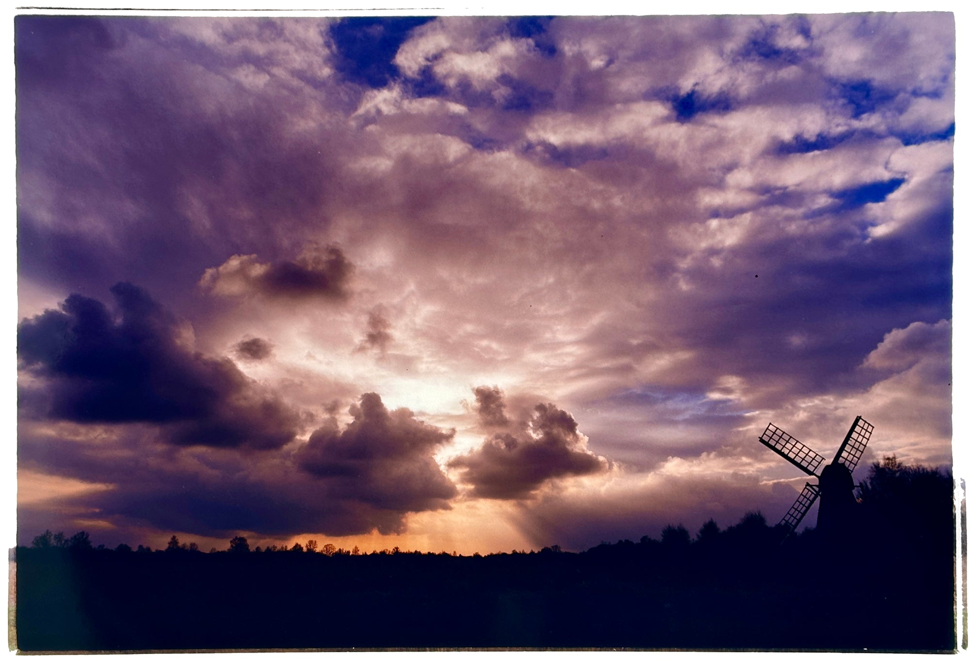 Photograph by Richard Heeps. A silhouette of a tree lined fen flat land with a windpump sitting on the right hand side. The sky is cloudy and bathed in golden dusk light.