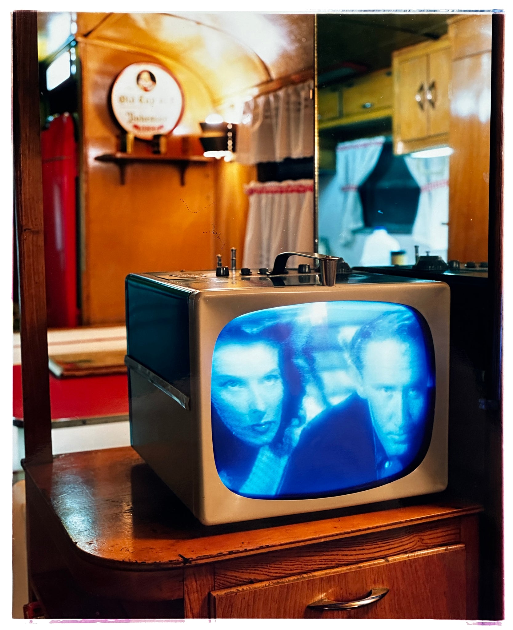 Photograph by Richard Heeps. A retro small brown television sits on a wooden table displaying a scene from a black and white film, Without Love. The image shows as blue and white. In the background of the television is a warm wood-panelled light.