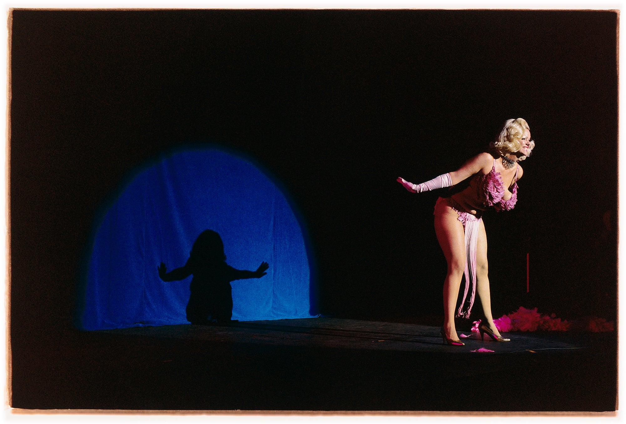 Photograh by Richard Heeps. A Burlesque dancer with a pink fluffy bikini bows on a black stage, in the background is a blue circle containing her silhouette.
