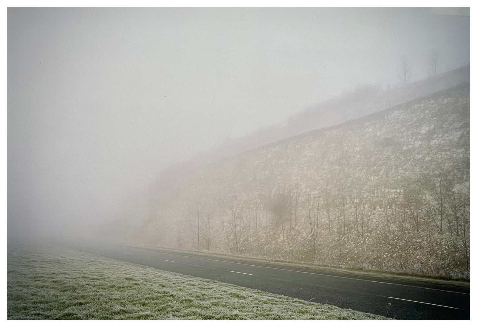 Photograph by Richard Heeps.  An empty road with an embankment on one side and grass on the other, all subdued by fog.