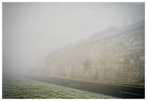 Photograph by Richard Heeps.  An empty road with an embankment on one side and grass on the other, all subdued by fog.