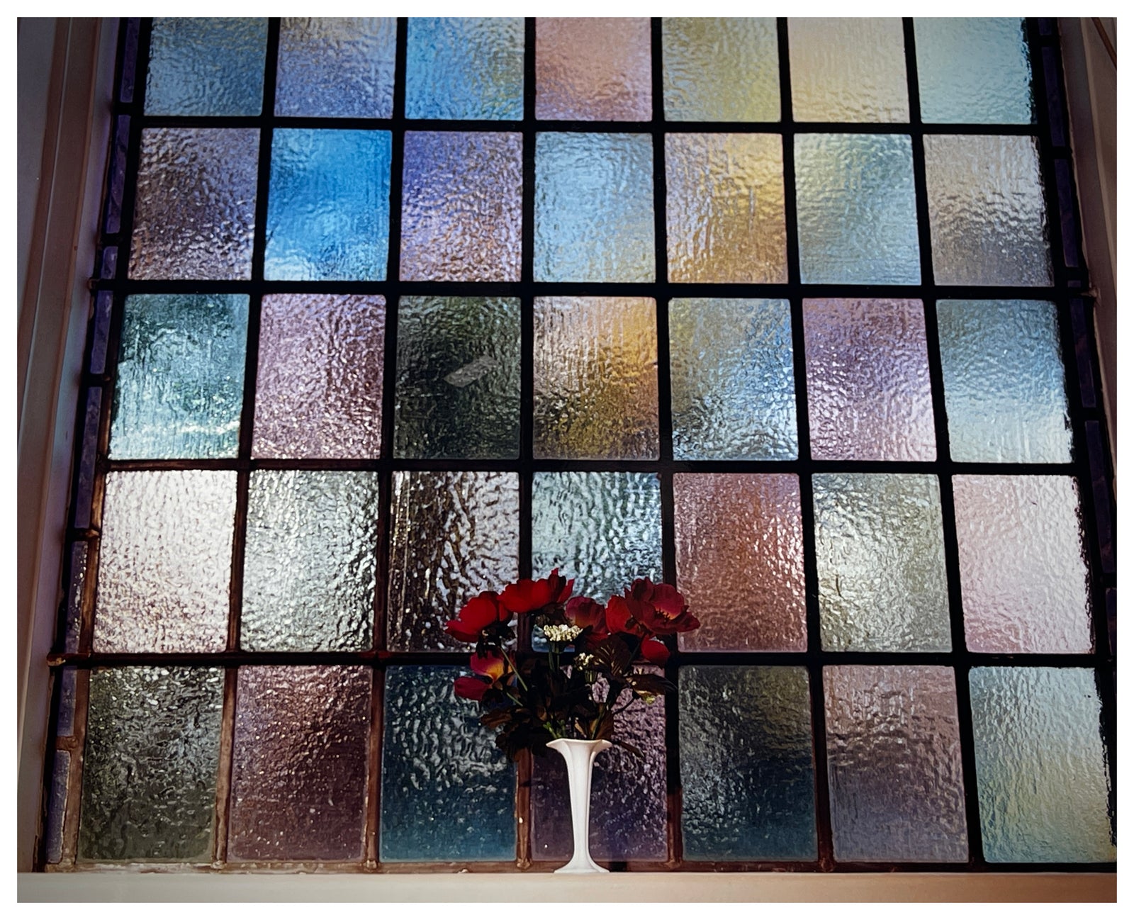 Photograph by Richard Heeps.   A small bunch of red flowers sit in a white vase in front of a coloured glass window.