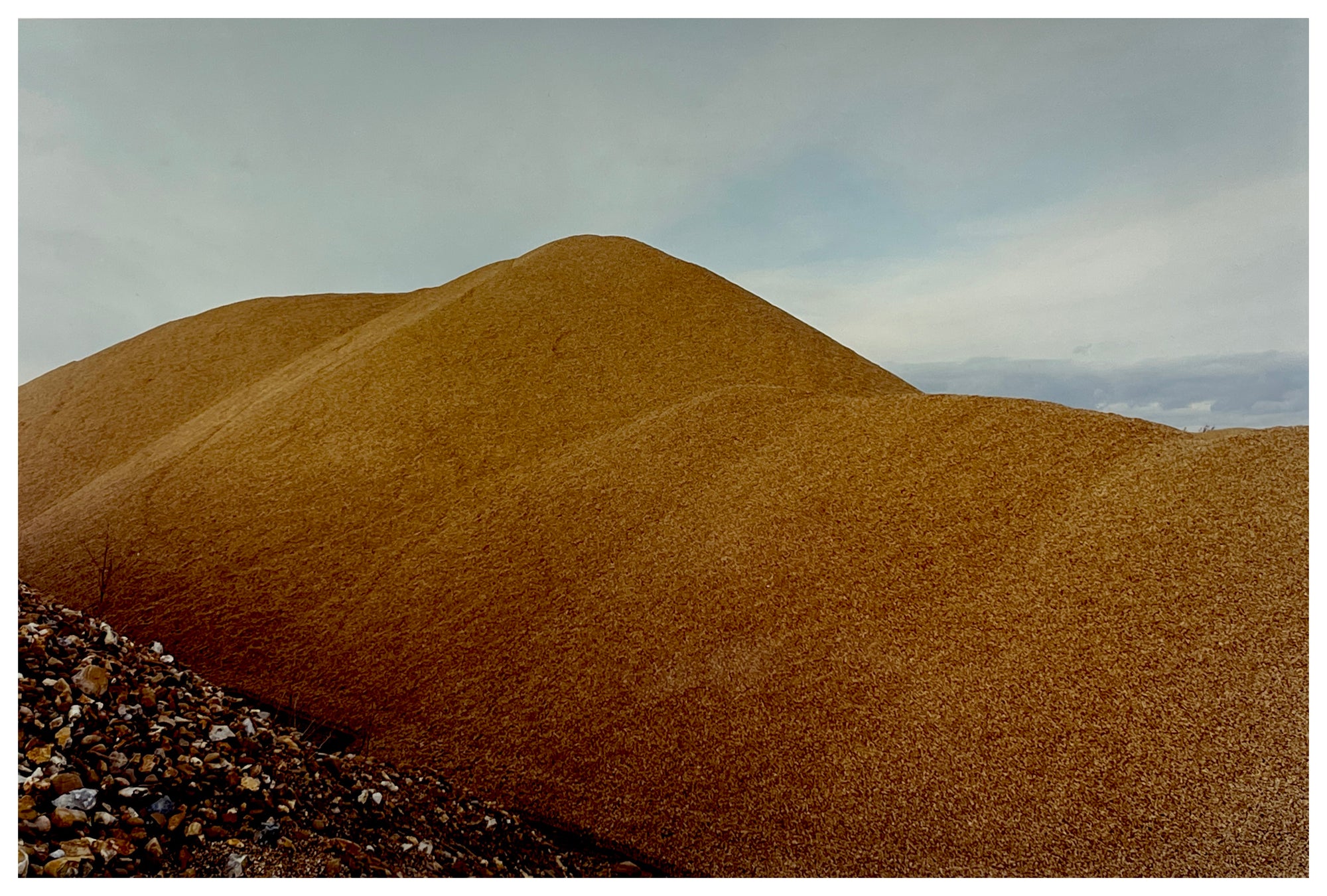 Photograph by Richard Heeps.  A golden pile of aggregate is piled up set against a cloudy sky.