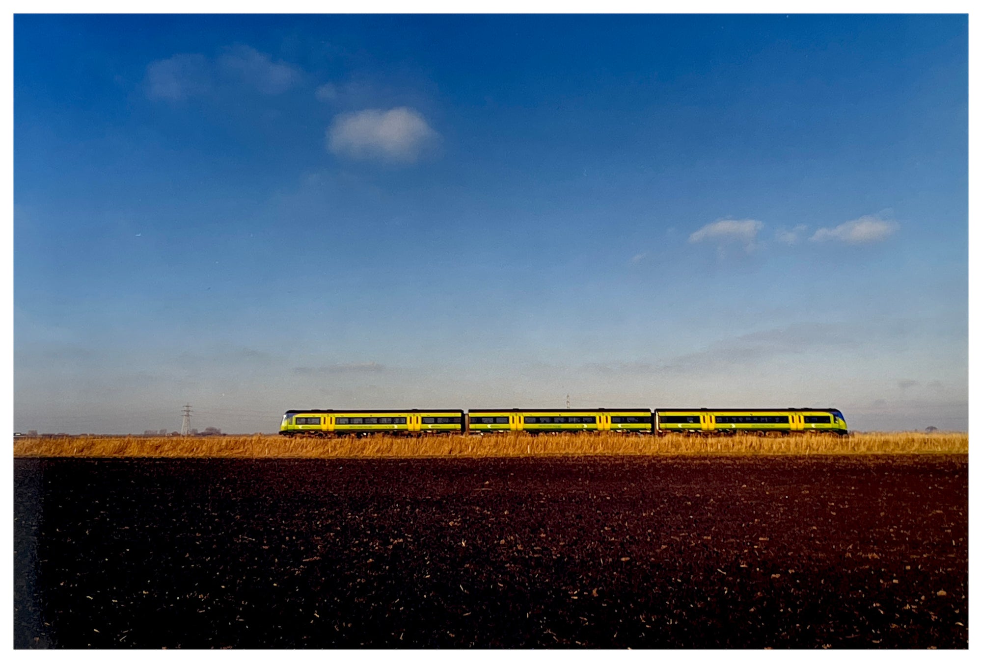 Photograph by Richard Heeps. This photograph has a yellow train travelling behind a fen embankment. A blue sky sits behind.