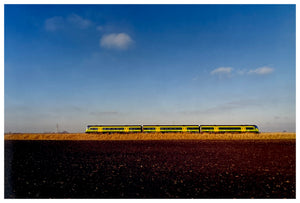 Photograph by Richard Heeps. This photograph has a yellow train travelling behind a fen embankment. A blue sky sits behind.