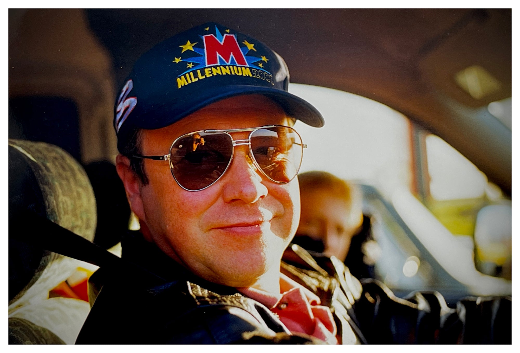 Photograph by Richard Heeps.  A smiling driver sits with brown sunglasses and a Millennium baseball hat.