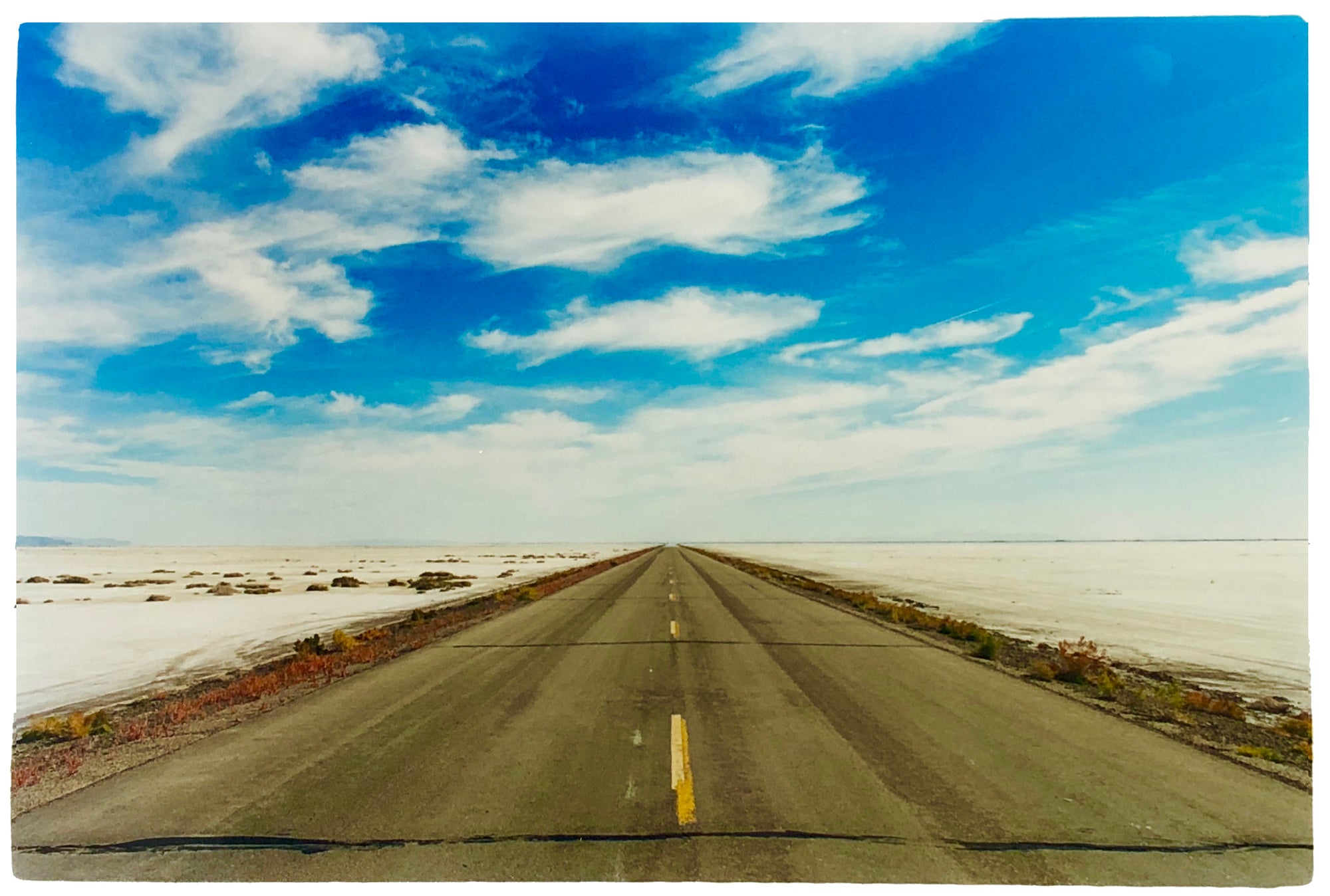 Photograph by Richard Heeps.  In the centre of the empty approach road going off into the distance, blue skies and white clouds fill the sky and either side of the road the flat salt plains.