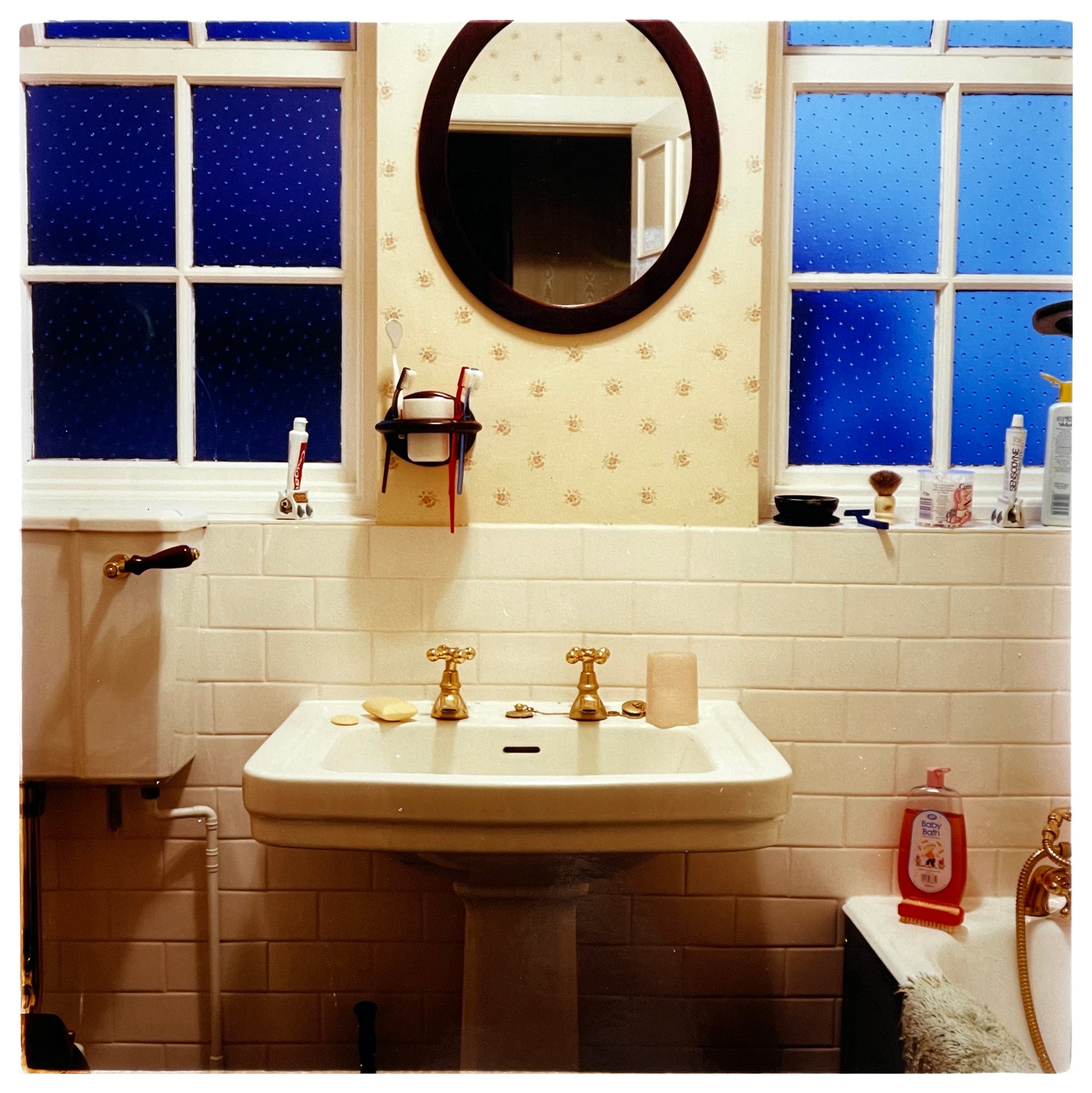 Photograph by Richard Heeps.  A basin, wth gold taps and an oval mirror over the top. There are frosted windows either side of the mirror.  