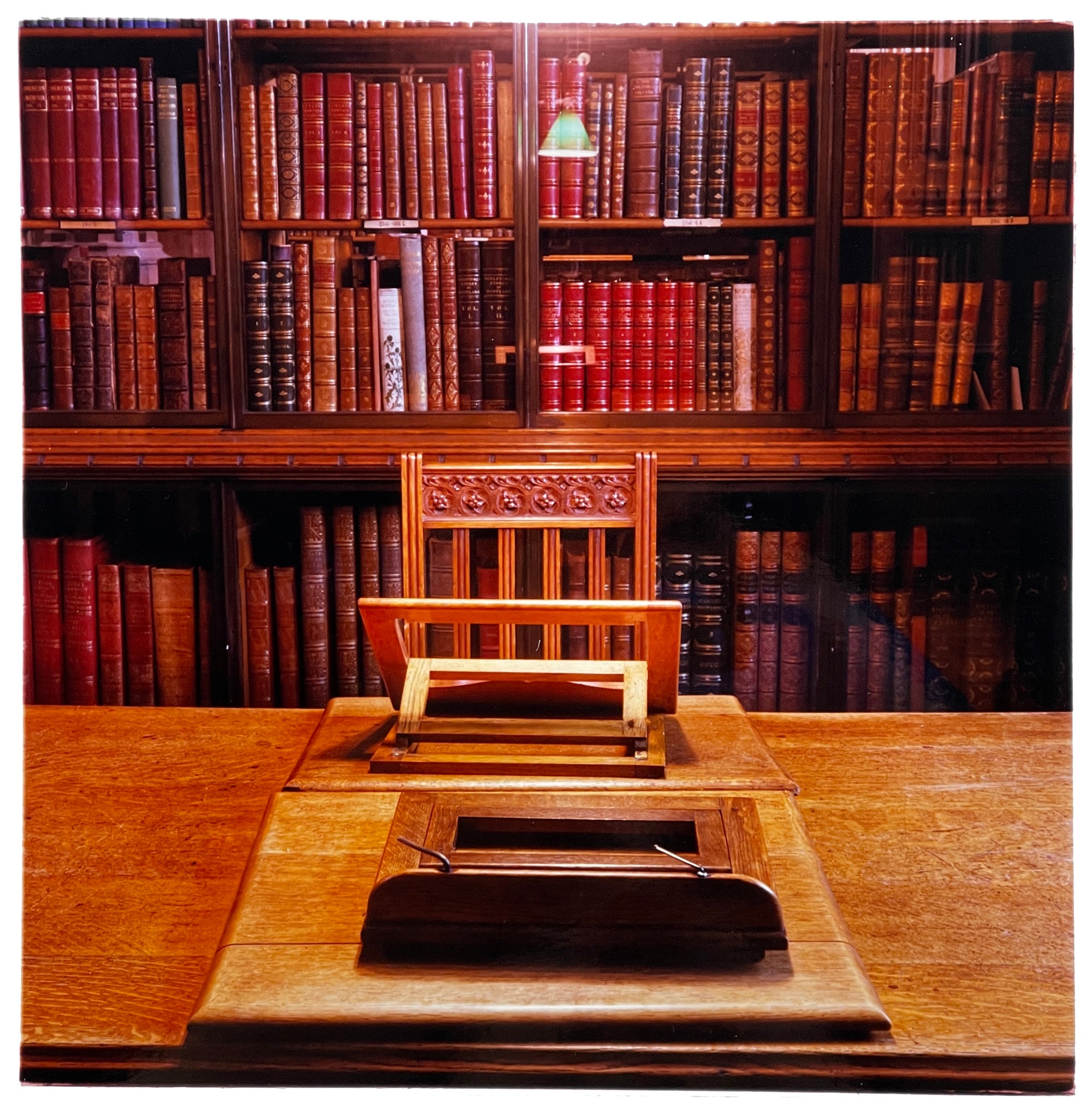 Photograph by Richard Heeps.  In the library with the books on the shelf behind and in foreground a wooden table with a chair.  The table has two easels lined up with the chair, the easel nearest the chair is open.
