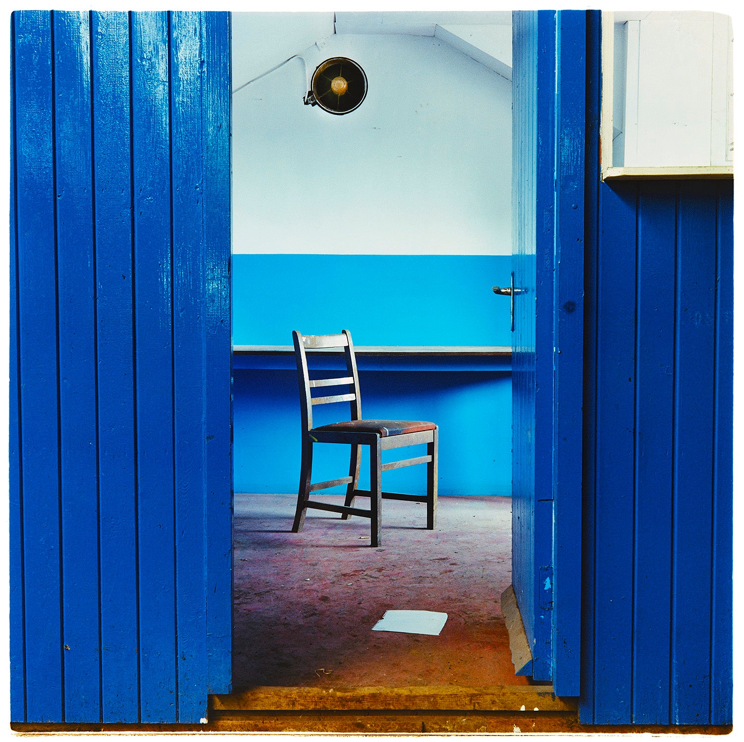 A photograph of chair which sits in a derelict factory in a colour block blue painted room, the perspective creates depth.