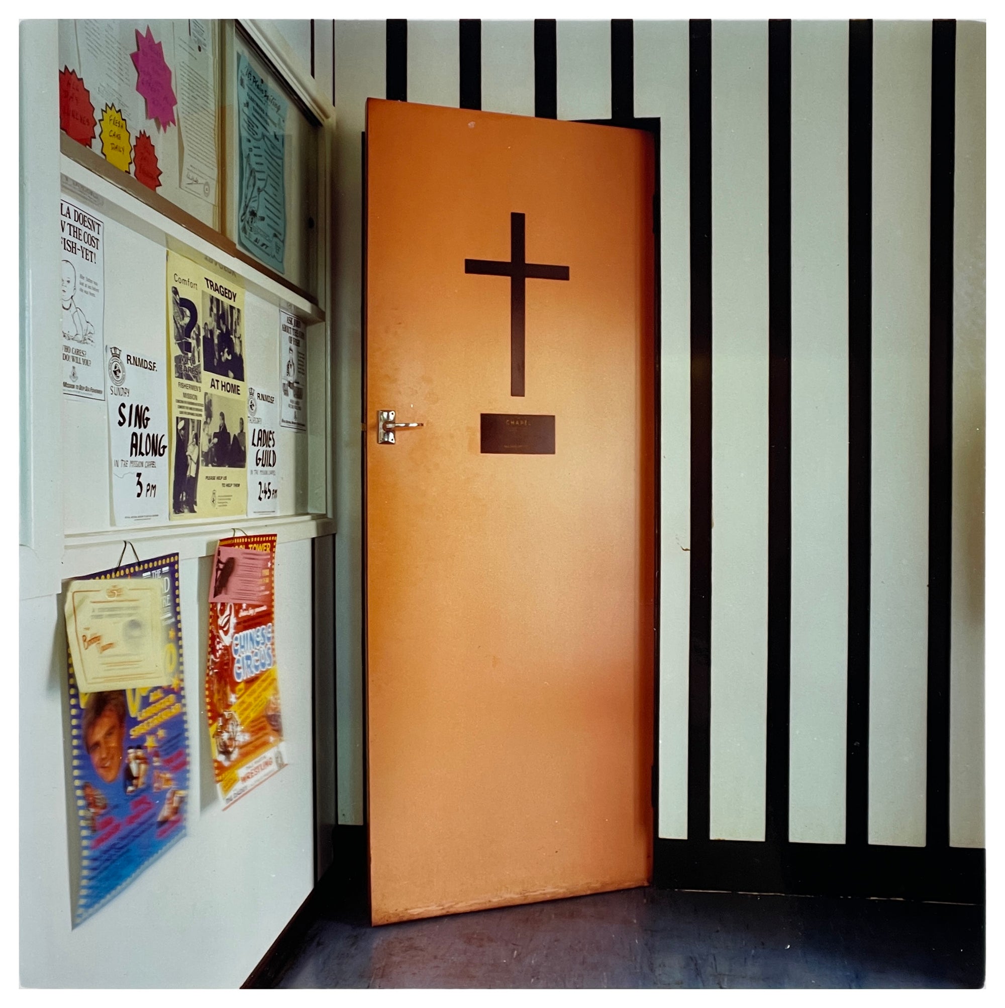 A photograph by Richard Heeps depicting a wooden door with a cross on it sitting slightly ajar set in a black and white striped wall. On the left hand side sits local community posters.