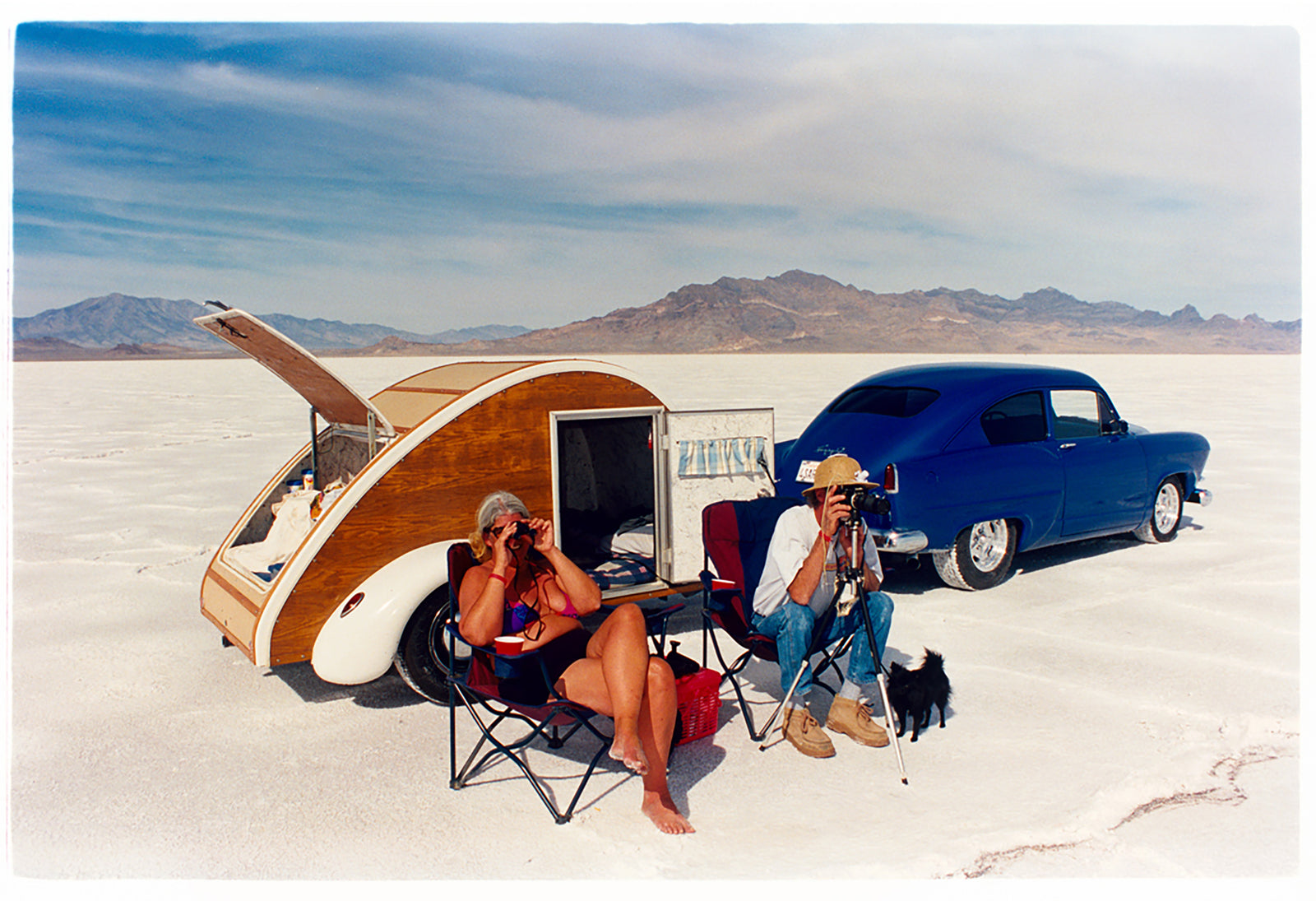'Christine's '52 Henry J & teardrop' was captured in Bonneville Salt Flats, Utah, the iconic home of speed. This photograph shows the mountains in the distance meeting and contrasting with the flatness of the salt pan, whilst a pair of retro spectators look out at the speeding cars in the distance.