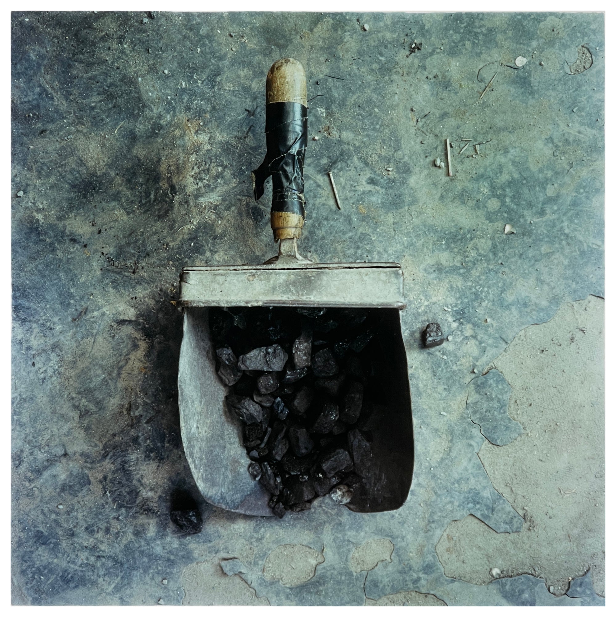 A photograph of a battered coal scuttle on the floor filled with coal, with its wooden handle taped. The coal scuttle sits on grey broken flooring. Photographer Richard Heeps